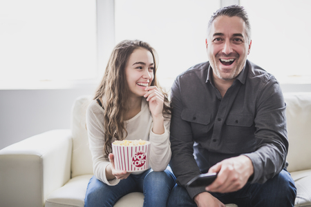 Portrait of a young man and daughter watching TV while eating popcorn on the sofaの写真素材