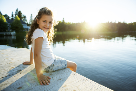 Cute child girl sitting on a wooden platform by the lake.の写真素材