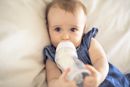 Pretty baby girl drinks water from bottle lying on bedの写真素材