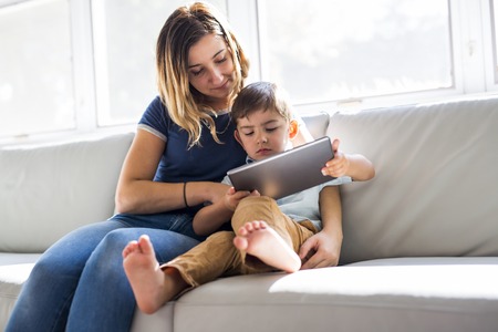 Little boy with digital tablet sitting on sofa, on home interior background with momの写真素材