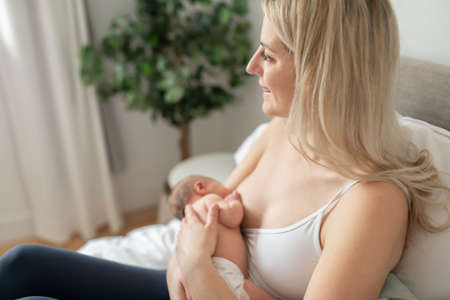 woman in her bedroom on the white cushions, smiling and breastfeed her baby.