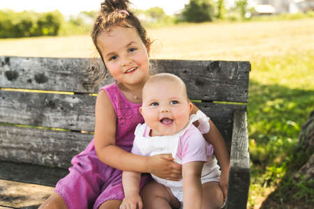Happy adorable chubby baby girl sitting on a bench with her daughter sisterの写真素材