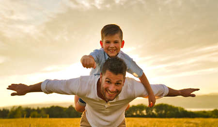 happy family of father and child on field at the sunset having fun flying on the shoulder