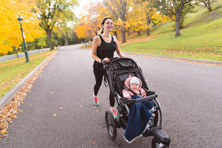 Beautiful young mother with her daughter in jogging stroller running outside in autumn nature