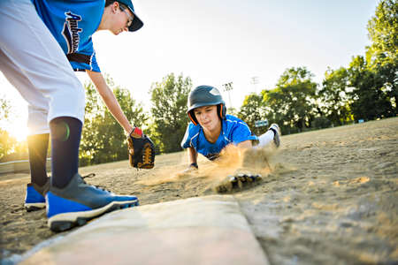 Group of two baseball players play together on the playground. On of them slide on the goal