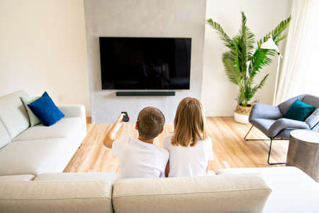 A two family brother and sister holding remote control and watching TV show. Back viewの写真素材
