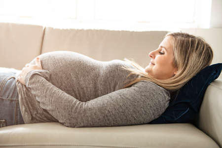 A happy pregnant woman lying on sofa at homeの写真素材