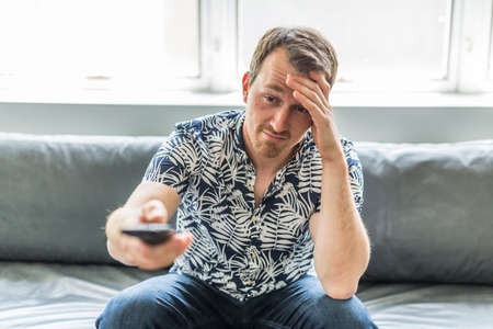 Bored man watching tv and zapping sitting on a couch at homeの写真素材