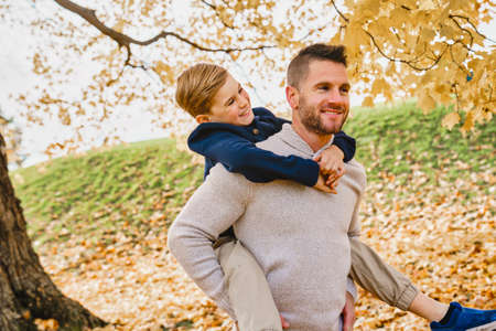 Happy family father and child boy in the autumn leaf fall in park holding his child on his backの写真素材