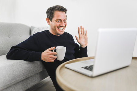 European man holding coffee while working on his laptop in bright living room.