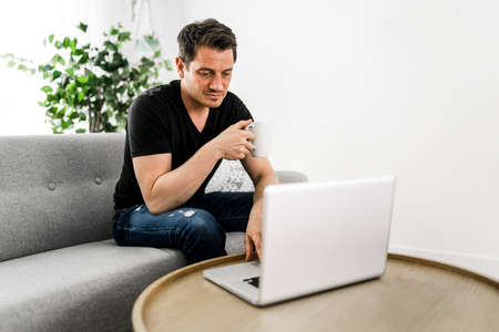 European man holding coffee while working on his laptop in bright living room.