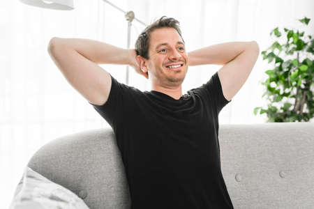 Relaxing man and dreaming on sofa at home with arm behind his head