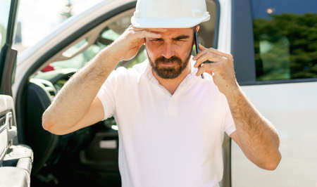 Man engineer builder wearing a white hard hat, shirt in front of his pickup using cellphone and seem...
