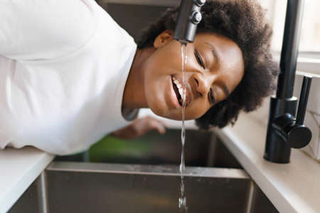 black woman drinking water directly from a kitchen faucetの写真素材