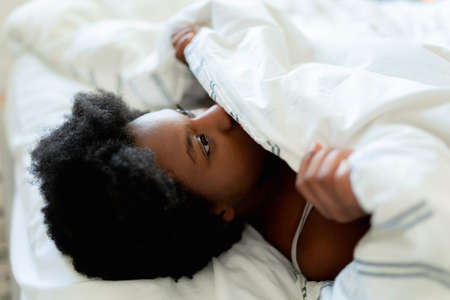 beautiful young African woman in tank top lying in bed cover face with sheetの写真素材