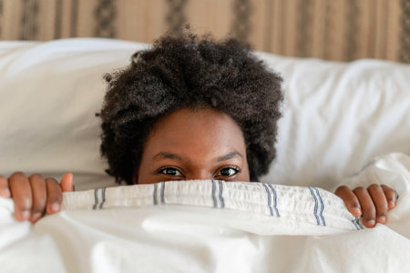 beautiful young African woman in tank top lying in bed cover face with sheet