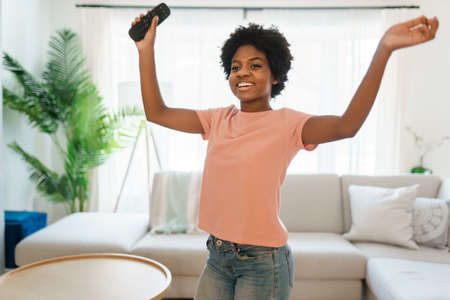 African woman shake head raise hands holds remote control in the living roomの写真素材