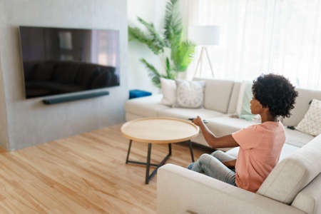 Cheerful cute woman sitting on sofa in bright living room changing tv channelの写真素材