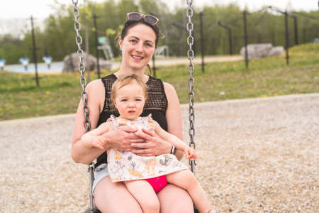 Cute baby girl playing on outdoor playground with mother on swingの写真素材