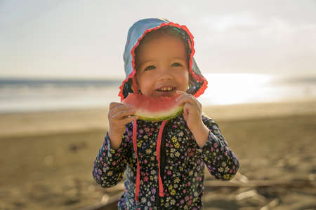 little adorable girl 2 years joyfully on the beach in sunset eating watermelonの写真素材