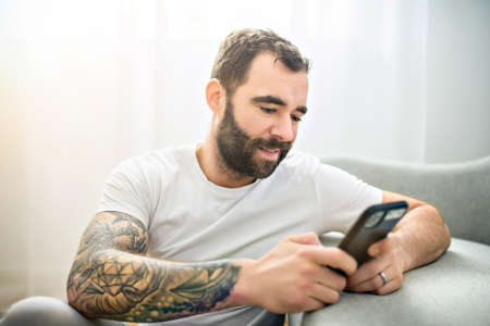 portrait of casual young man with beard and tattoo and white shirt sit on the sofa at home with cellphoneの写真素材
