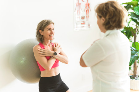 A Elderly Physiotherapist working with sporty patient in clinic. woman do wall squats with fit ball