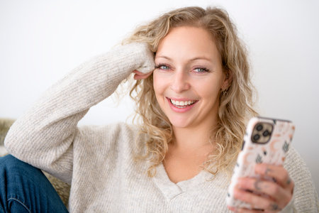 Happy attractive blonde woman at home close to a window with cellphoneの写真素材