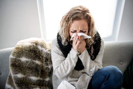 upset young woman sitting on sofa covered with blanket freezing blowing running nose got fever caught cold sneezing in tissue, sick girl having influenza symptoms coughing at home, flu conceptの写真素材