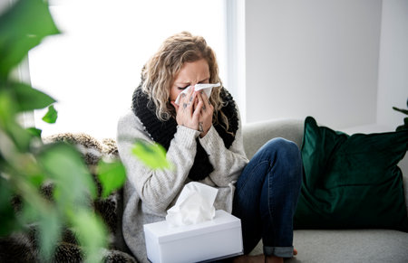 upset young woman sitting on sofa covered with blanket freezing blowing running nose got fever...