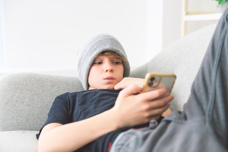 young boy lay on the couch at home watching cellphone wearing toqueの写真素材