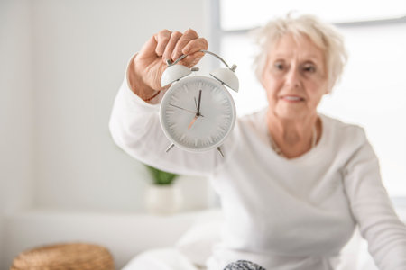 Smiling senior woman resting on bed holding clockの写真素材