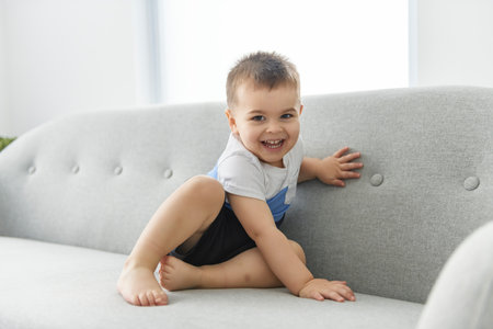 little boy 2 years old sits on bottom of comfortable sofa at homeの写真素材