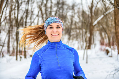 Female runner jogging in cold winter forest wearing warm sporty running clothing