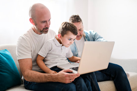 Happy two man couple with adopted child sit on sofa at home using laptop