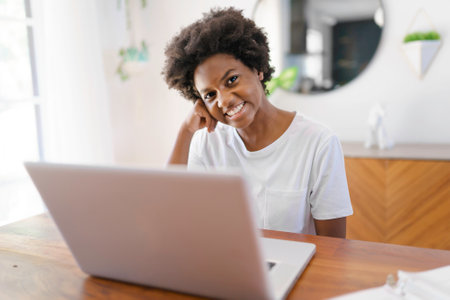 Smiling young African female entrepreneur working online with a laptop while sitting at her kitchen...