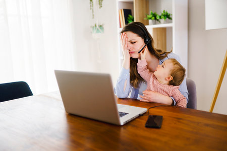 sad and frustrated mother in kitchen home office with computer and her daugherの写真素材