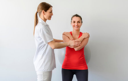 Physiotherapist helping woman do wall squats with fit ball. Young female patient doing back exercise using fitball in physio room of modern clinicの写真素材
