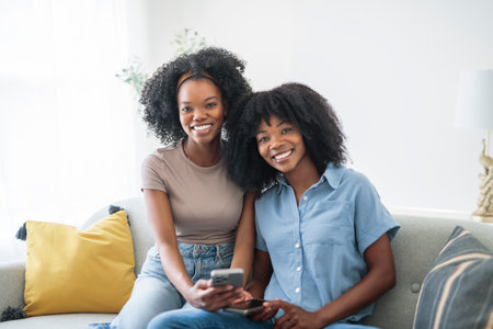 black mother and daughter using cellphone on sofa