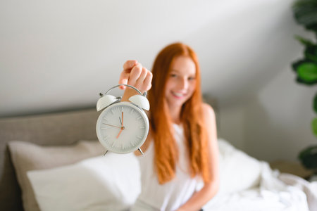 Young redhead woman on bedroom at home with clock close toの写真素材