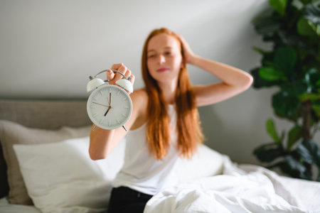 Young redhead woman on bedroom at home with clock close to