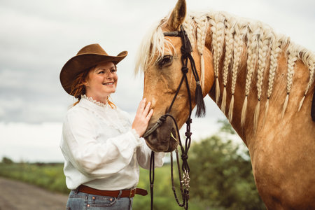 A woman is wearing a cowboy hat while standing next to a horse in an open fieldの写真素材