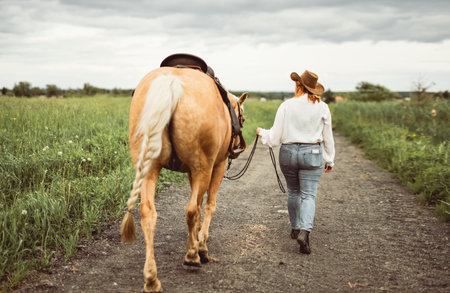 A woman is wearing a cowboy hat while walking beside a horse on a dirt roadの写真素材