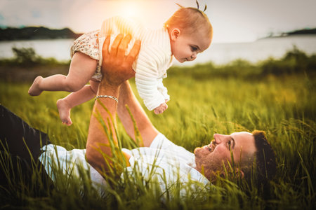 father and baby child girl at the sunset lay on the grass on the waterscape view from backの写真素材