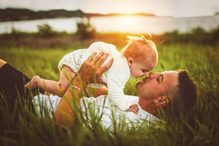father and baby child girl at the sunset lay on the grass on the waterscape view from backの写真素材