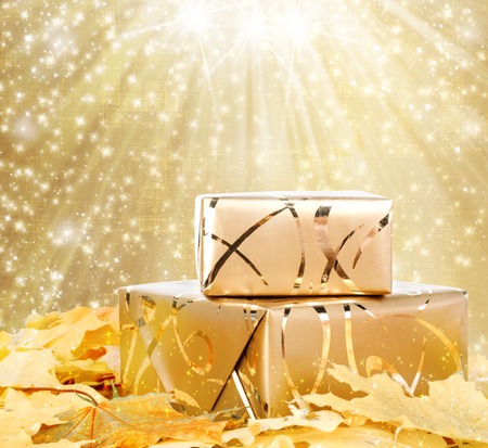 Gift box in gold wrapping paper with autumn leaves on the abstract glowing backgroundの写真素材