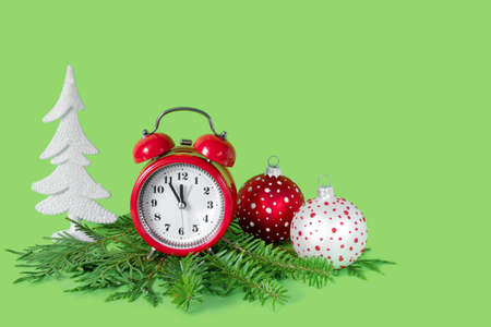 Red and white Christmas balls, alarm clock and tree branches on green paper background. 5 minutes before the New Year. Minimal celebration concept with copy space.の写真素材