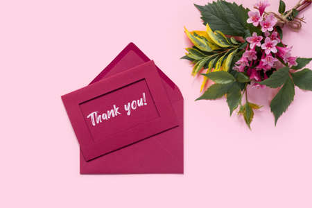 Beautiful bouquet of flowers of delicate weigela on pink background. Envelope and letter with the inscription Thank you! Minimal concept backdrop. Flat lay top view with copy space.の写真素材