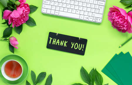 Remote home office on green desk. Workspace with keyboard and pencils. Bouquet of beautiful delicate pink peonies and cup of hot tea for holiday. Thank you lettering on the black board. Appreciation for the service.の写真素材