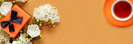 Gift box with white hydrangea and rose flowers on orange background. Cup of hot tea. Conceptual postcard for the holiday. Flat lay, top view concept with copy space for text.の写真素材