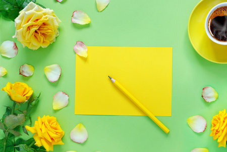 Bouquet of beautiful yellow roses and cup of hot fresh aromatic coffee on green paper background. Notebook sheet and pencil for your text. Flat lay, top view, copy space concept.の写真素材